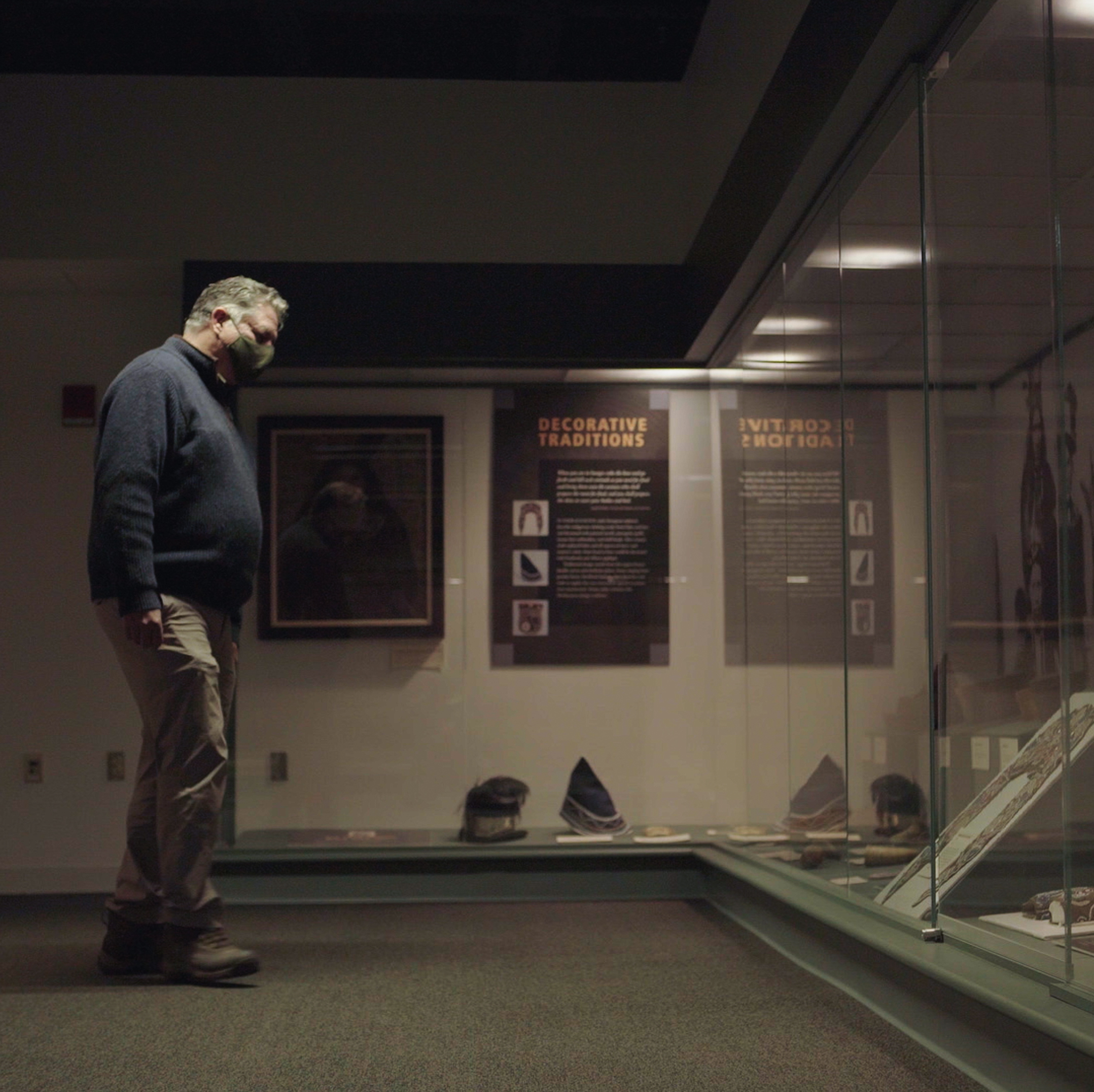 Photograph of James Eric Francis Senior standing in a darkened museum, looking at Penobscot clothing items displayed in glass cases.